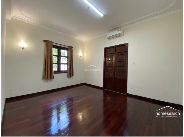 This bedroom features rich, dark polished hardwood flooring that contrasts elegantly with the soft cream-colored walls and crown molding. The room is equipped with a wall-mounted AC unit, warm ambient wall sconces, and a window with security bars and neutral curtains, providing a well-maintained and classic atmosphere.