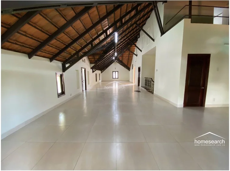 This expansive attic-style room features a dramatic vaulted ceiling with exposed dark wood beams and a large, open floor plan finished with light-colored tile flooring. The space is brightened by multiple windows and a central staircase, offering a versatile area that appears to be in good condition.