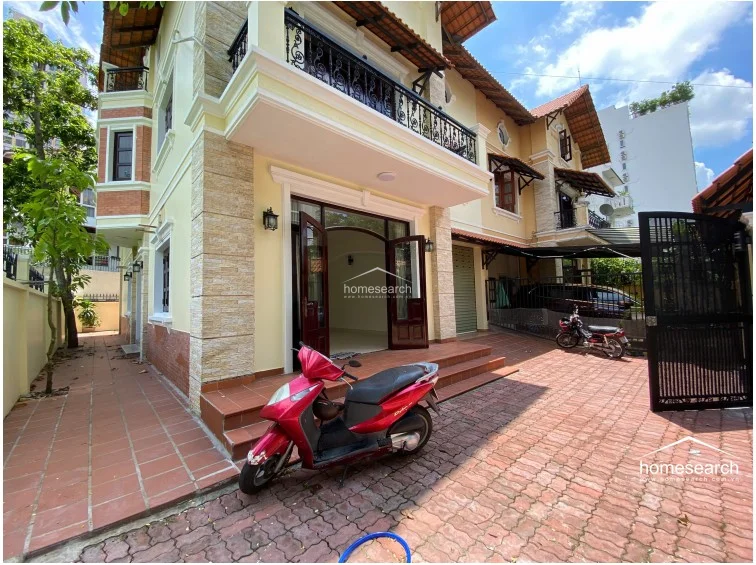 The exterior of this multi-story villa showcases a warm yellow facade with stone accents and a terracotta tiled roof. The property features a paved driveway, a gated entrance, and a balcony with decorative wrought-iron railings, reflecting a well-maintained Mediterranean-inspired architectural style.
