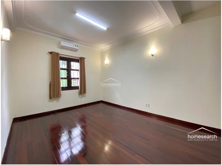 This bedroom mirrors the design of the previous one, showcasing dark, high-gloss hardwood floors and cream-colored walls with decorative ceiling trim. It is outfitted with a functional AC unit, wall-mounted lighting fixtures, and a window with security bars, maintaining a consistent and polished aesthetic throughout the home.