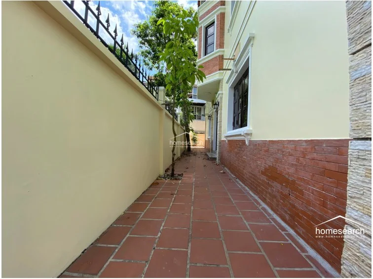 This exterior side passage features terracotta-style floor tiles and a high perimeter wall topped with decorative iron spikes for security. The building facade showcases a blend of light-colored stucco and a lower brick-veneer base, with a young tree providing a touch of greenery in the narrow space.