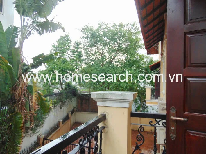 This balcony area is accessed via a dark wood door and features a decorative black wrought-iron railing. It offers an elevated view of the surrounding greenery and the property's lower-level exterior, with the building's tiled roof overhang providing shade.