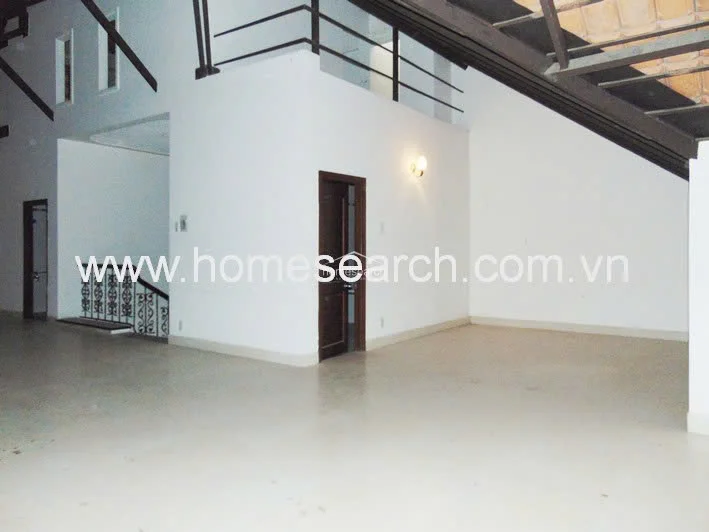 This spacious, open-plan interior features high, vaulted ceilings with exposed structural beams and a mezzanine level overlooking the main floor. The space is finished with clean white walls and neutral tile flooring, complemented by a dark wood door and a decorative wrought-iron staircase railing that adds a touch of classic architectural detail.