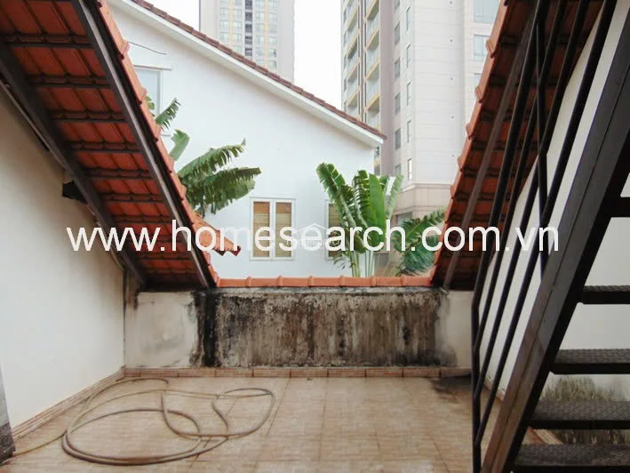 This outdoor terrace area features terracotta-tiled flooring and an open-air design framed by sloping rooflines and a metal staircase. While it offers a unique architectural aesthetic and views of neighboring buildings and greenery, the low wall shows significant signs of weathering and moisture damage, indicating a need for maintenance.
