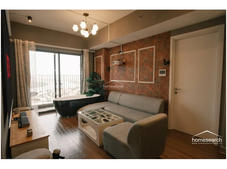 This open-concept living area features a modern, unconventional layout with a freestanding black soaking tub placed directly by the floor-to-ceiling balcony windows. The space is finished with wood-look flooring, a textured orange accent wall with white molding, and a grey sofa, all illuminated by a decorative chandelier and recessed ceiling lights.