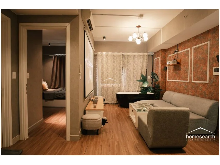 The living area flows seamlessly into a dining space and entryway, with a projector screen serving as a primary entertainment feature. The neutral grey walls and warm wood tones create a cohesive aesthetic, while the wall-mounted AC unit and minimalist furniture maintain a clean, uncluttered feel.