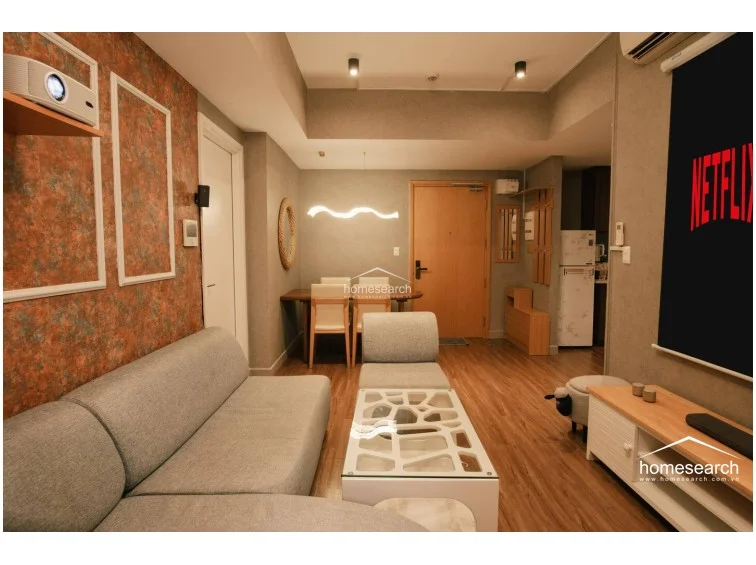 This wide view captures the entire flow of the unit, from the entrance door and dining area to the living room and kitchen corner. The layout is highly efficient, utilizing a projector for media to save space, while the light-colored wooden cabinetry and functional entryway storage reflect a well-organized and modern living environment.