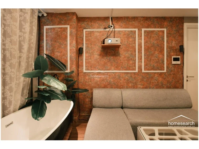 This unique living area features a bold, textured orange-toned wallpaper with white trim accents and a modern grey sectional sofa. A freestanding soaking tub is positioned near the window, creating a spa-like atmosphere, while a projector mounted on the wall provides entertainment options. The space is finished with warm wood-look flooring and sheer lace curtains that soften the natural light.