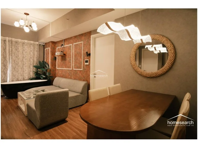 The dining area is defined by a sleek, modern wavy light fixture and a circular rope-framed mirror, providing a stylish transition from the living zone. A wooden dining table with upholstered chairs anchors the space, maintaining the warm and inviting atmosphere established throughout the apartment.