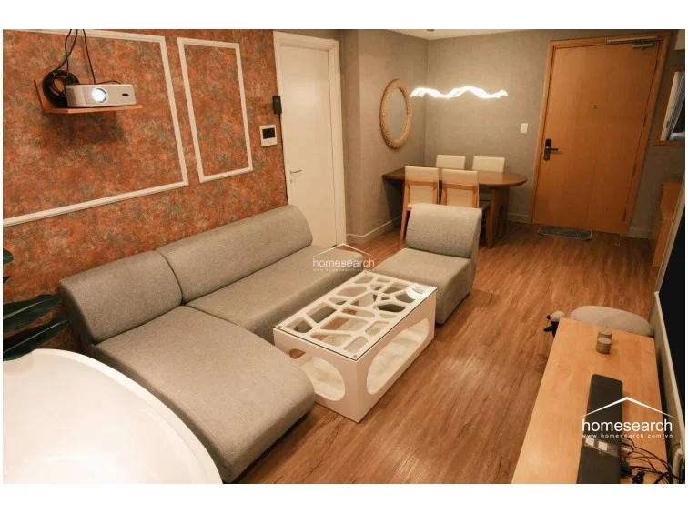The living room layout integrates a dining area near the entrance, featuring a wooden table with four upholstered chairs and a decorative rope-framed mirror. The space maintains a cohesive aesthetic with wood-look flooring throughout and a contemporary white glass-top coffee table. A projector setup and a wall-mounted digital control panel highlight the unit's modern, tech-forward functionality.