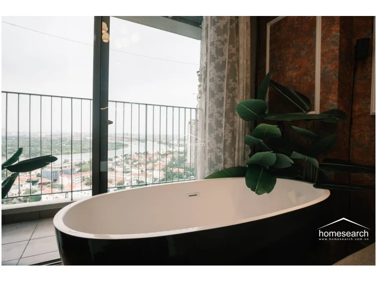 This image showcases a striking freestanding soaking tub positioned directly against a floor-to-ceiling window, offering expansive views of the city and river. The area is accented by a large decorative plant, which adds a touch of nature to the sophisticated, modern interior. The combination of the sleek tub and the panoramic backdrop creates a luxurious, relaxing focal point within the room.