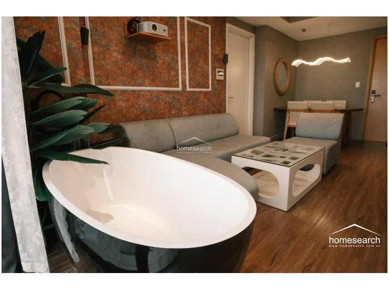 This angle highlights the unique placement of the soaking tub, which offers a scenic view through the large glass balcony doors. The room is well-maintained, featuring a contemporary glass-topped coffee table and a large artificial plant that adds a touch of greenery to the sophisticated interior.
