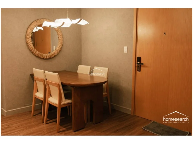 The dining area is efficiently arranged near the main entrance, featuring a warm-toned wooden dining table and four cream-colored chairs. The walls are finished in a neutral, textured grey, complemented by a unique, sculptural light fixture and a circular rope-accented mirror. The wood-look flooring flows seamlessly from this space into the rest of the unit, maintaining a consistent and polished appearance.