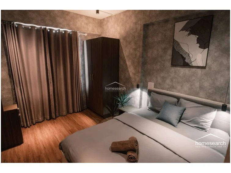 This bedroom features a calming, monochromatic color scheme with textured grey walls and matching bedding. A tall, dark wood wardrobe provides essential storage, while heavy floor-length curtains allow for complete light control. Modern pendant lights hang above the bed, and a piece of abstract wall art adds a sophisticated touch to the well-maintained, contemporary sleeping space.