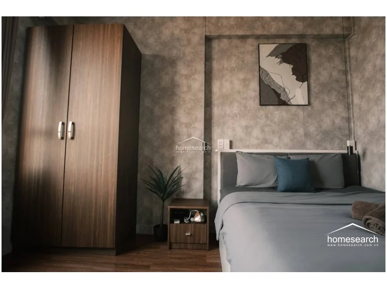 This compact bedroom features a modern, moody aesthetic with textured grey walls and warm wood-look flooring. It is furnished with a double bed, a tall dark-wood wardrobe, and a matching bedside table, creating a cohesive and minimalist living space.
