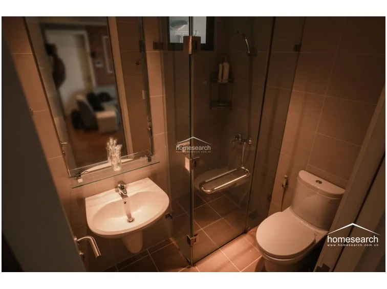 The bathroom layout is efficient, utilizing a corner shower enclosure to maximize the available floor space. The room is finished with warm-toned tiling and features a wall-mounted mirror above the sink, providing a clean and functional design.