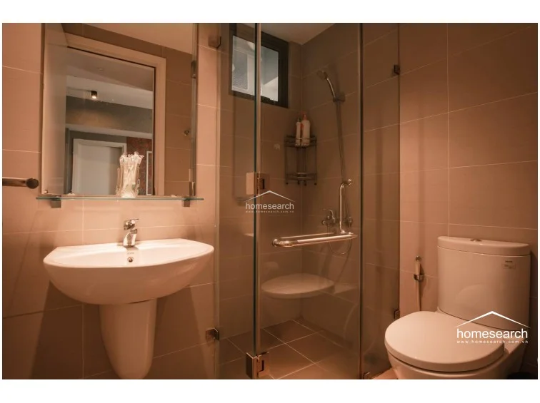 This compact bathroom features a modern, neutral aesthetic with light beige wall tiles and matching floor tiles. It includes a pedestal sink, a glass-enclosed walk-in shower with a built-in seat, and a standard white toilet, all maintained in good condition.