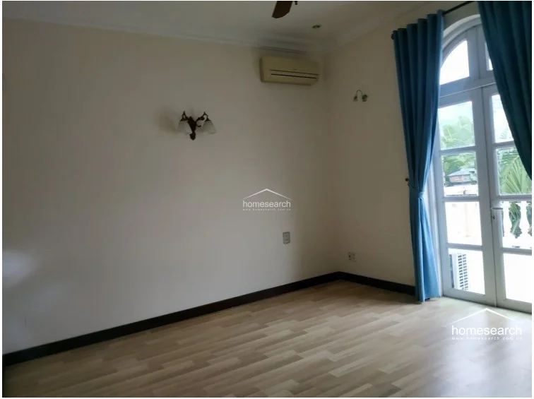This bedroom features rich, dark wood flooring and neutral walls, creating a warm and classic atmosphere. It is equipped with a wall-mounted AC unit, blue curtains for privacy, and a traditional wall sconce for ambient lighting.