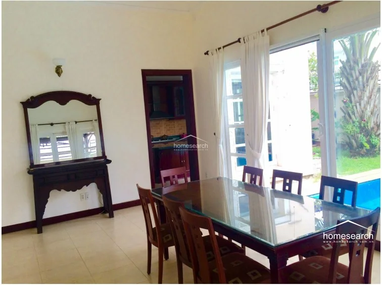 This living room features a bright, open-plan layout with light-colored tile flooring and a grand archway leading to a staircase with ornate iron railings. The space is furnished with traditional wooden seating featuring vibrant red cushions, creating a formal and welcoming environment.
