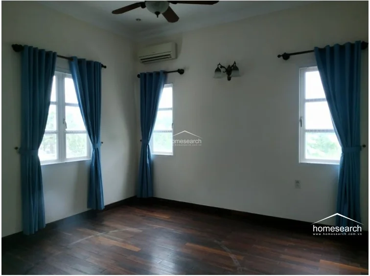 This room is finished with warm-toned hardwood flooring and features a large sliding glass door that provides ample natural light and a view of the greenery outside. The space is equipped with a wall-mounted air conditioning unit and is framed by simple white curtains.