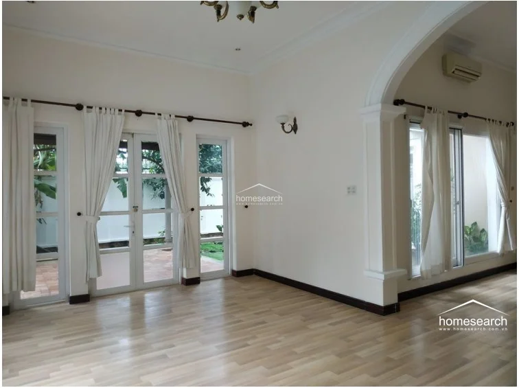 This spacious living area features light wood-look flooring, white walls, and a decorative ceiling with a classic chandelier. A prominent structural column is wrapped in green artificial turf, and the room connects to an outdoor space via an arched glass door.