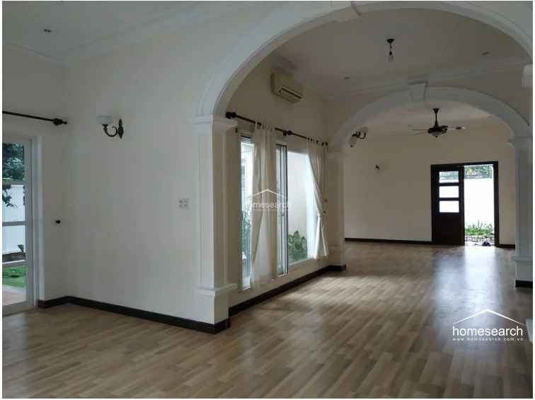 This expansive room includes light wood-look flooring and a built-in wooden shelving unit, offering functional storage space. The layout is open and airy, featuring an arched doorway and a central column, with the entire space appearing well-maintained.