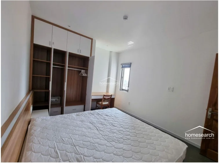This bedroom is efficiently designed with a large, multi-section built-in wardrobe that incorporates both hanging space and shelving, along with a built-in desk by the window. The room is finished with white walls and grey floor tiles, maintaining a neutral and tidy appearance that maximizes the available square footage.