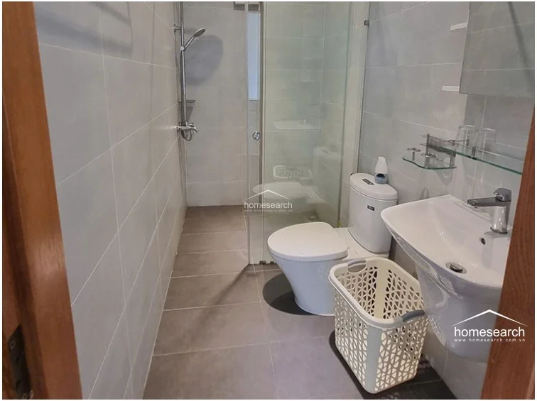 The bathroom is compact and functional, featuring a glass-enclosed shower stall and light-colored wall tiles that contrast with the darker grey floor tiles. It is equipped with standard white porcelain fixtures, including a wall-mounted sink and a toilet, and appears to be in well-maintained, clean condition.