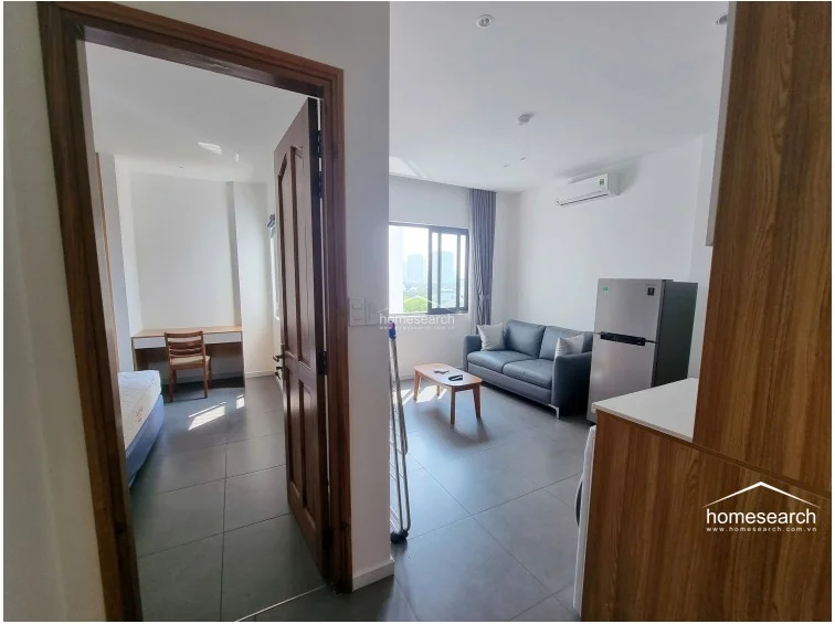 This view from the doorway captures the flow between the bedroom and the open-plan living area, both featuring consistent grey tile flooring and white walls. The bedroom includes a built-in desk and a simple bed frame, while the living area is equipped with a wall-mounted AC unit and a grey sofa, all bathed in natural light from the window.