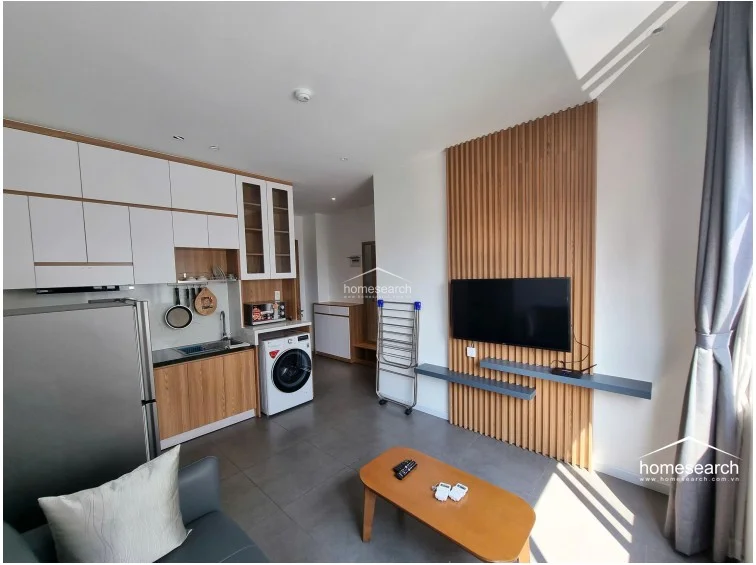 This living area features a modern kitchenette with white upper cabinets and wood-grain lower cabinets, complemented by a stainless steel refrigerator and a front-loading washing machine. A decorative vertical wood-slat feature wall serves as the focal point for the television, while the space maintains a clean, minimalist aesthetic with grey floor tiles.