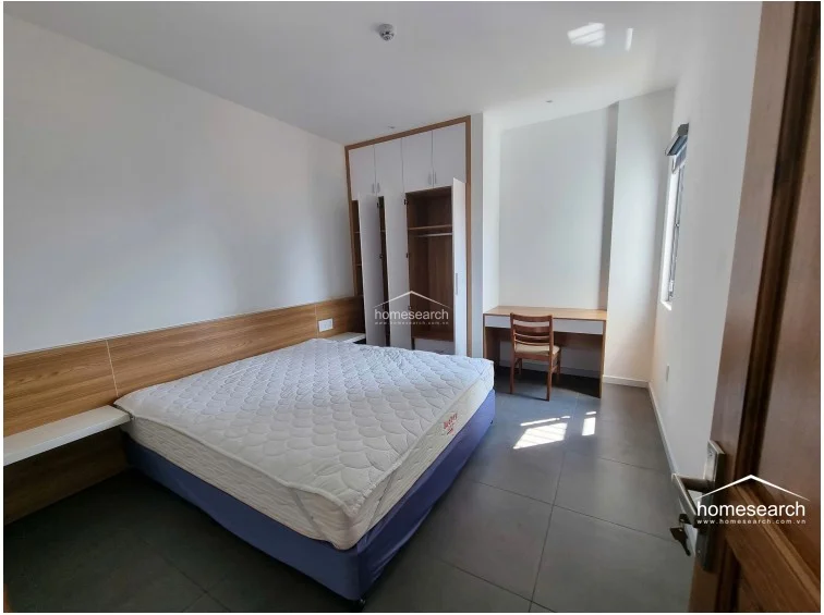 This angle highlights the bedroom's functional layout, showing the connection between the sleeping area, the built-in storage, and the dedicated workspace. The room is well-lit by a single window and features a consistent, modern color palette of white and natural wood tones.