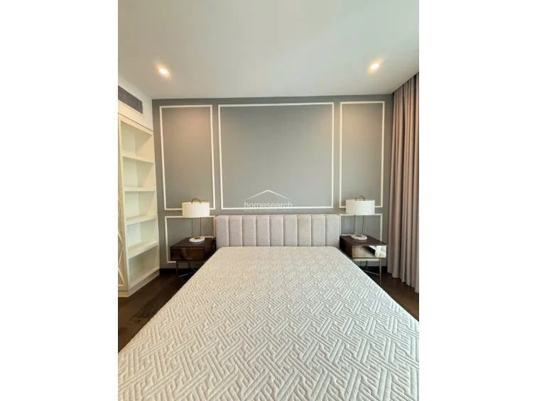 This elegant bedroom features a plush upholstered headboard set against a grey accent wall with classic white molding. The room includes dark wood flooring, built-in shelving, and mirrored nightstands, creating a refined and well-maintained atmosphere.