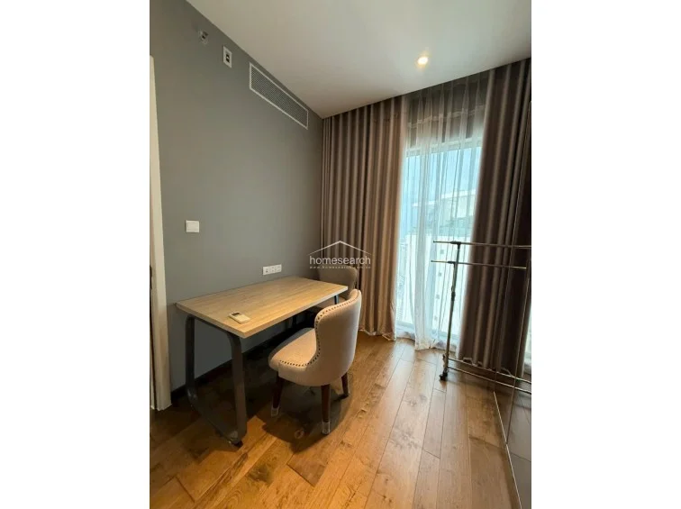 This compact study or workspace features warm wood-tone flooring and a minimalist desk setup with matching upholstered chairs. The room is illuminated by natural light from a window draped in floor-to-ceiling taupe curtains, and it includes a built-in ceiling AC unit for climate control.