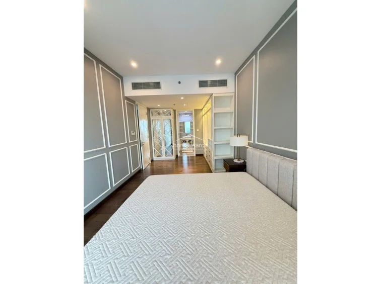 This master bedroom showcases an elegant design with grey walls accented by classic white wainscoting and warm wood flooring. It includes a plush upholstered bed, built-in open shelving for storage, and a seamless transition into a walk-in closet and en-suite bathroom area.