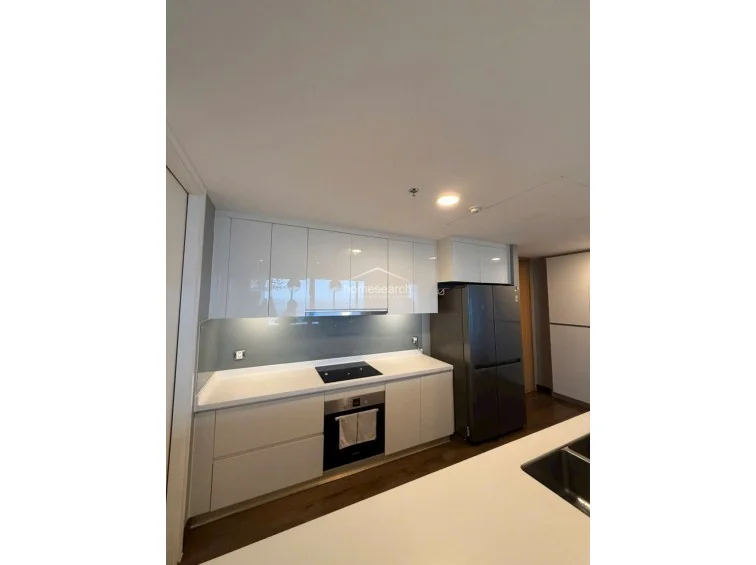 This close-up of the kitchen highlights the high-end, minimalist design, featuring glossy white upper and lower cabinets and a clean white countertop. The area is equipped with a modern induction cooktop, a built-in oven, and a large, dark-toned refrigerator, all maintained in excellent condition.