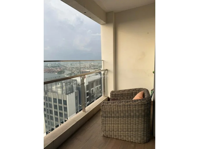 This balcony offers a sleek, modern aesthetic with a glass railing that maximizes the expansive city and river views. It features warm-toned wood-look flooring and a comfortable wicker armchair, creating a serene spot for relaxation.