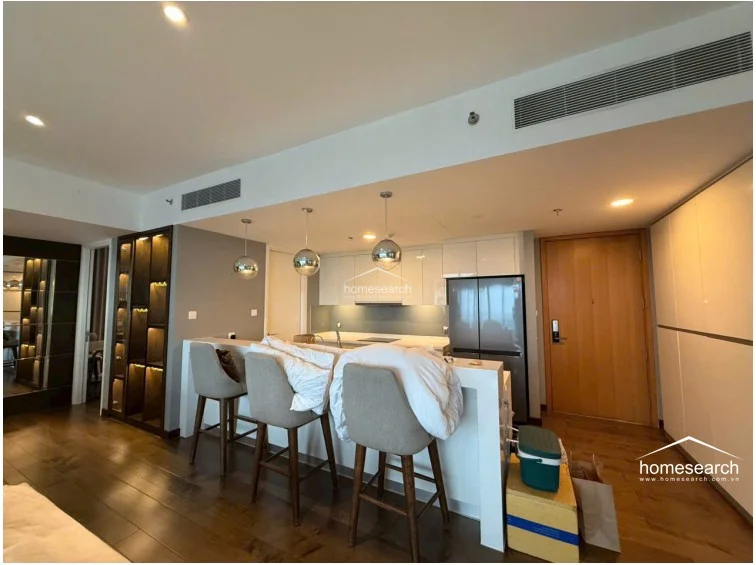 This view of the kitchen and entryway highlights a contemporary layout with white high-gloss cabinetry, integrated appliances, and a breakfast bar illuminated by three spherical pendant lights. The area is seamlessly connected to the living space and features ample storage, including a full-height pantry and a decorative, backlit display cabinet.
