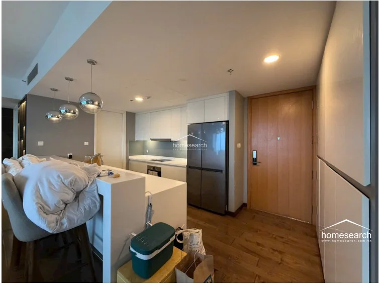 This open-concept kitchen and entryway area features warm wood flooring and a sleek, modern aesthetic with white cabinetry and a contrasting grey backsplash. The space is well-lit by recessed ceiling lights and stylish pendant lamps hanging over the breakfast bar, which serves as a functional transition to the living area.
