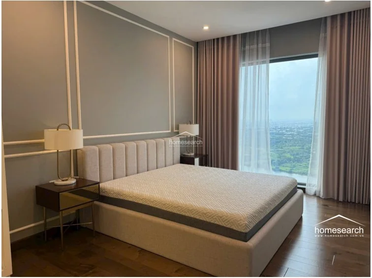 This elegant bedroom features a plush, tufted headboard and sophisticated wall molding, set against a backdrop of soft grey walls and rich wooden flooring. A large floor-to-ceiling window offers an expansive, scenic view, complemented by layered curtains that allow for adjustable natural light.
