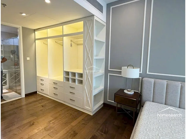 The bedroom includes a custom-built, open-concept wardrobe system with integrated lighting, providing both hanging space and dedicated shelving for organized storage. The room maintains a cohesive design with the adjacent bathroom visible through the doorway, showcasing a high level of maintenance and modern interior detailing.