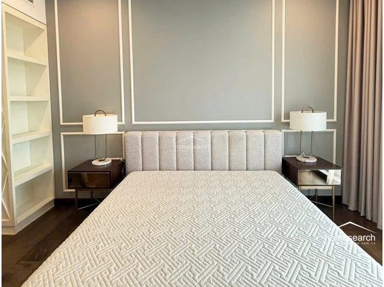This bedroom features a sophisticated design with light gray walls accented by classic white molding and a plush, tufted headboard. The space is well-appointed with matching bedside tables, elegant lamps, and a built-in floor-to-ceiling white shelving unit for organized storage.