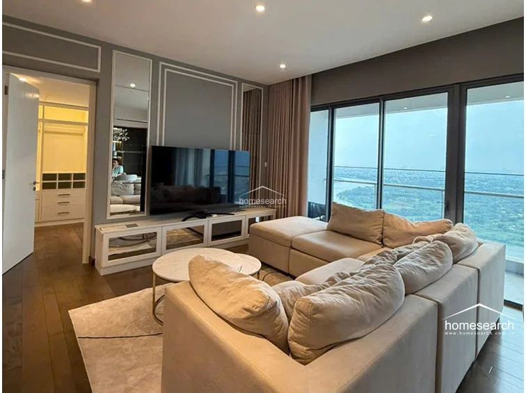 This modern living area features a spacious sectional sofa and a sleek entertainment unit with integrated mirror paneling. Large floor-to-ceiling windows provide expansive city and river views, while the room is finished with sophisticated wall molding and recessed ceiling lighting.