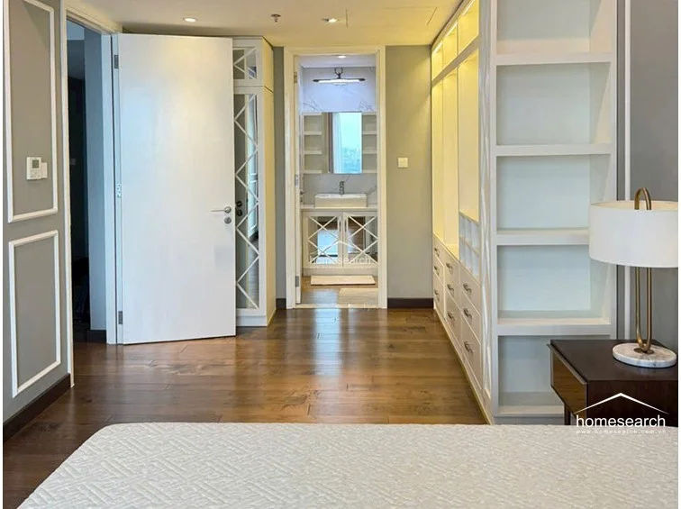 This bedroom layout flows directly into an en-suite bathroom, highlighting a seamless transition between living and private areas. The room is equipped with extensive custom cabinetry, including open shelving and closed drawers, and features high-quality dark wood flooring throughout.