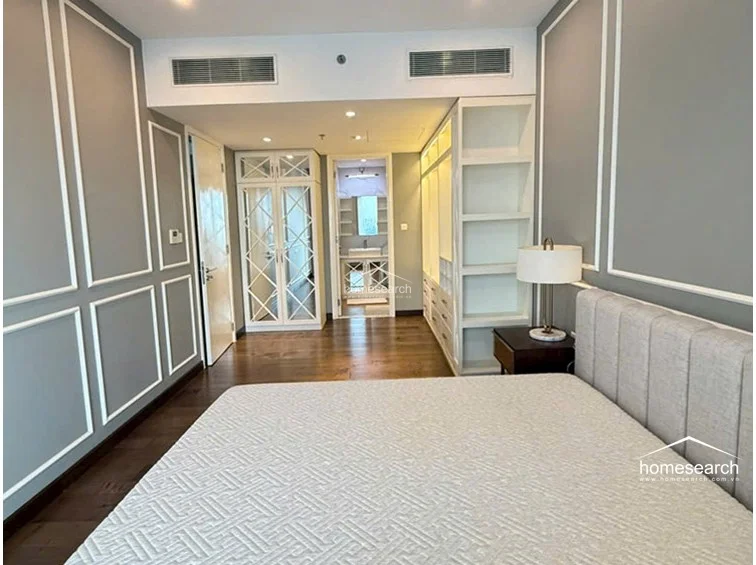 This bedroom features a sophisticated design with grey walls accented by white molding and warm-toned wood flooring. It includes a plush, tufted headboard, built-in open shelving, and a mirrored wardrobe that leads directly into a bright, modern bathroom.