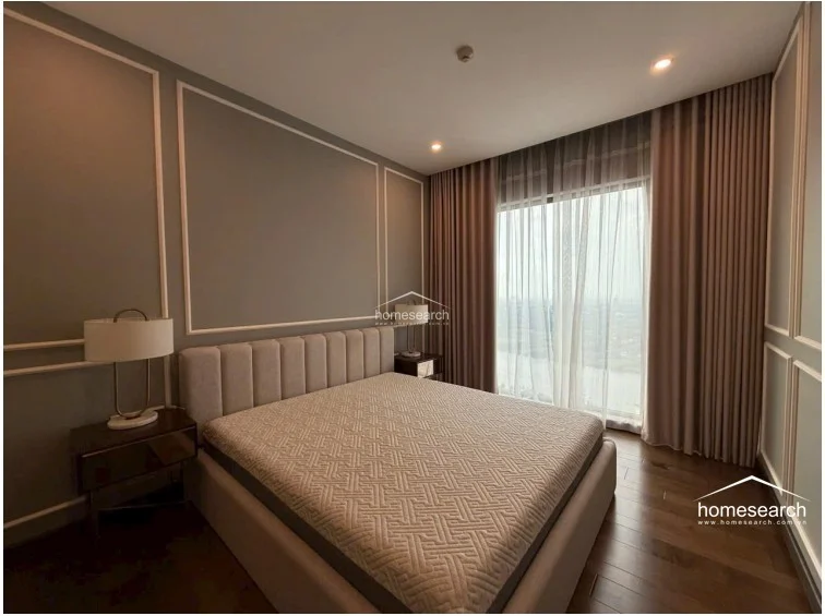 The bedroom is bathed in natural light from a large floor-to-ceiling window dressed in elegant floor-length curtains. The space is anchored by a contemporary upholstered bed and matching bedside tables, maintaining a clean, high-end aesthetic throughout.