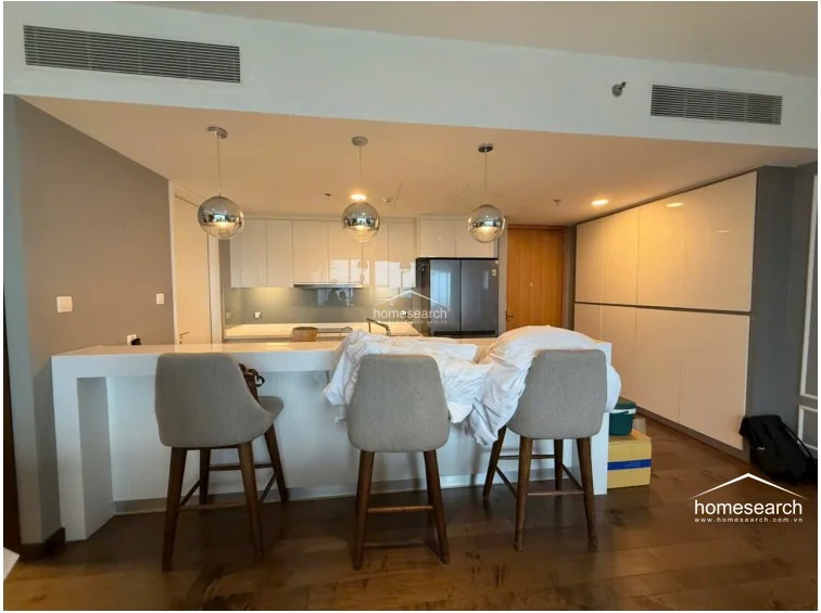 The kitchen is designed with a sleek, minimalist aesthetic, featuring high-gloss white cabinetry, a breakfast bar with three upholstered stools, and modern pendant lighting. The area is equipped with integrated appliances and a mirrored backsplash, all set against a warm wood floor that connects the kitchen to the rest of the open-plan living space.