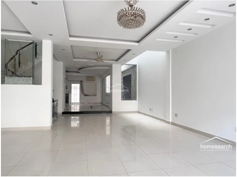This expansive open-plan living area features high ceilings with sophisticated tray lighting and a decorative crystal chandelier. The space is finished with polished light-colored floor tiles and white walls, creating a bright and airy atmosphere that flows seamlessly toward the rear of the home.