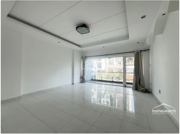 This spacious living area features a minimalist design with white walls, a tray ceiling with recessed lighting, and polished white tile flooring. Large sliding glass doors provide excellent natural light and direct access to a private balcony overlooking the neighborhood.