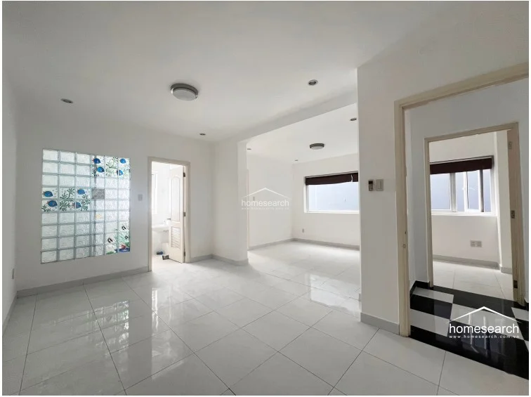 This interior space acts as a transition area, connecting multiple rooms through a clean, open layout with white walls and consistent white tile flooring. A unique glass block wall adds a decorative element, while a section of the floor features a striking black-and-white checkered tile pattern near the doorway.