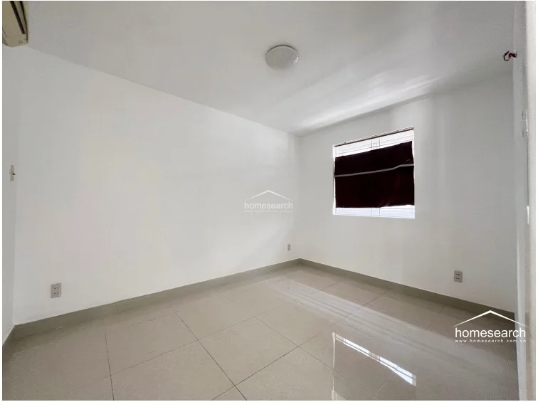 This bedroom is a simple, neutral space finished with white walls and light-colored tile flooring. It includes a single window with a dark roller blind for privacy and is pre-equipped with an AC unit mounted on the wall.